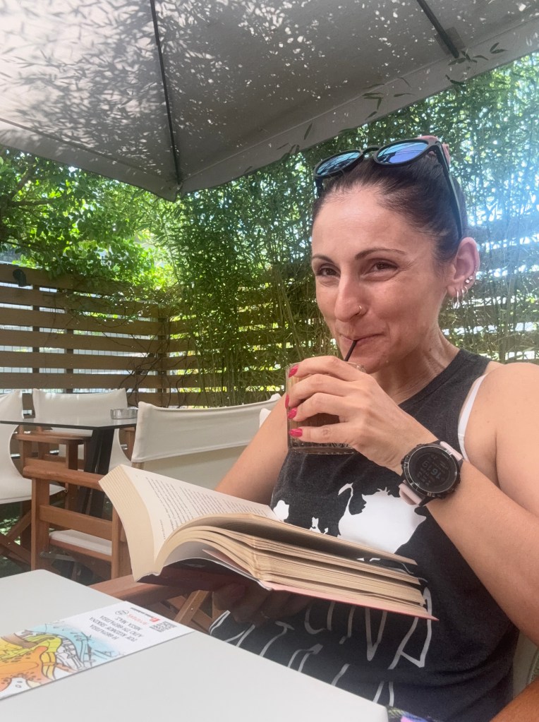 Γυναίκα κάθεται σε καφετέρια διαβάζοντας ένα βιβλίο και πίνει καφέ από ένα ποτήρι.